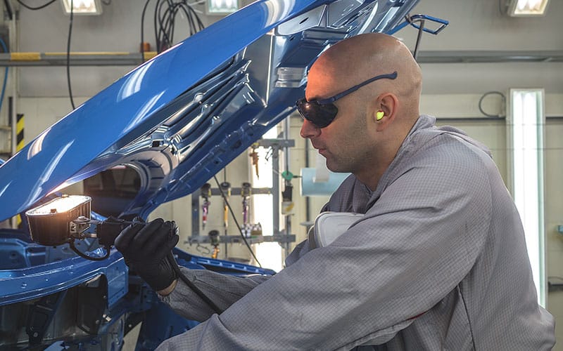 A worker in safety glasses and bright custom earplugs inspecting a car body under a shop light